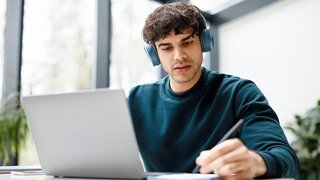 Remote studying. Concentrated european guy in headphones watching video course on laptop and taking notes