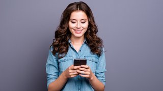 Content trendy cheerful nice cute adorable lovely attractive brunette girl with wavy hair in casual denim shirt, typing in phone, isolated over grey background