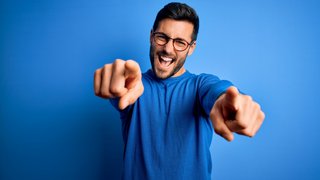 Young handsome man with beard wearing casual sweater and glasses over blue background pointing to you and the camera with fingers, smiling positive and cheerful