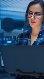 Portrait of a Smart Young Woman Wearing Glasses Holds Laptop. In the Background Technical Department Office with Specialists Working and Functional Data Server Racks